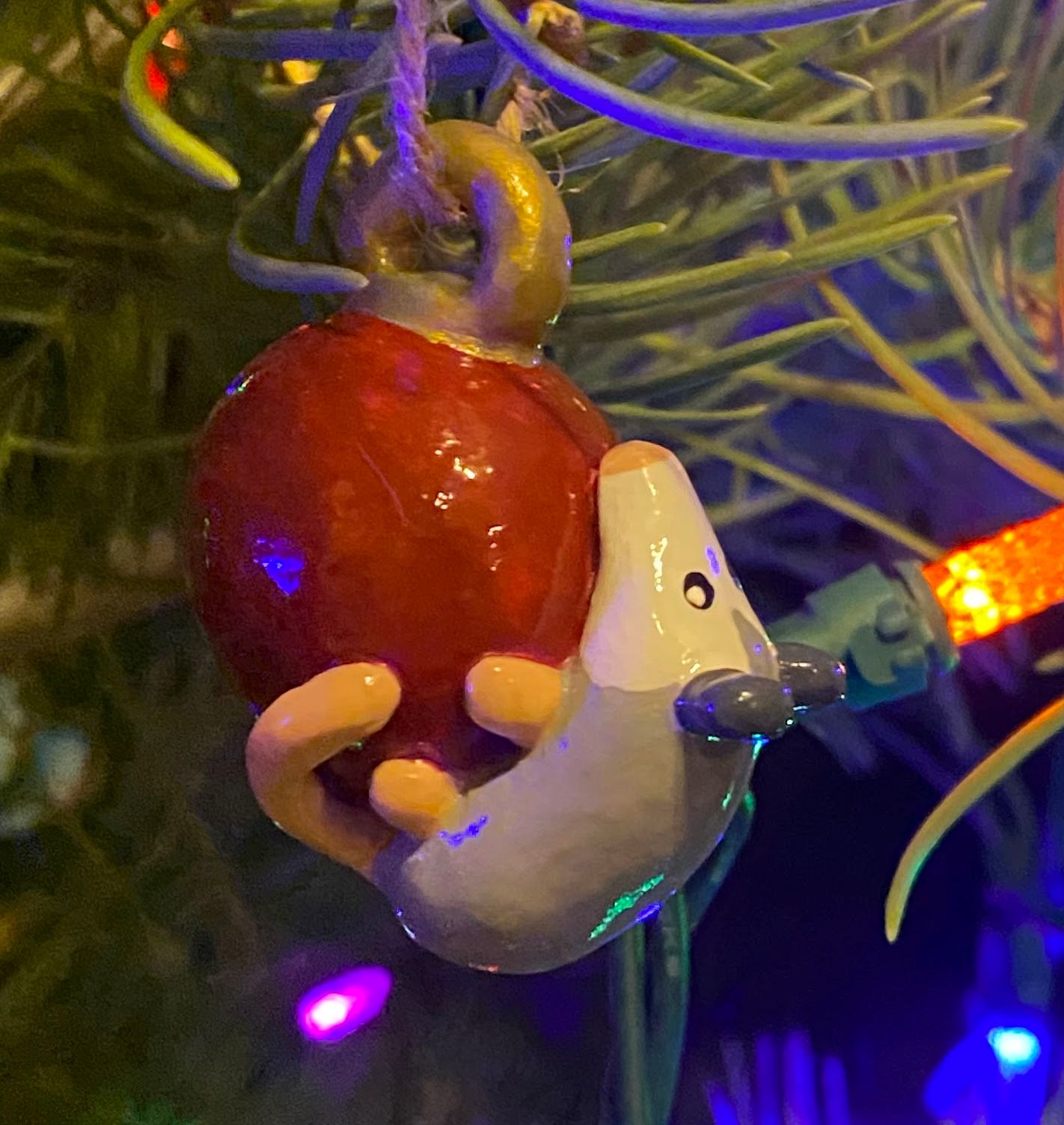 A possum hangs from a red christmas tree ball ornament. A christmas tree sits in the background